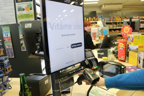 Detailní záběr na ruku zákazníka skenujícího produkt ručním skenerem u obrazovky samoobslužné pokladny.