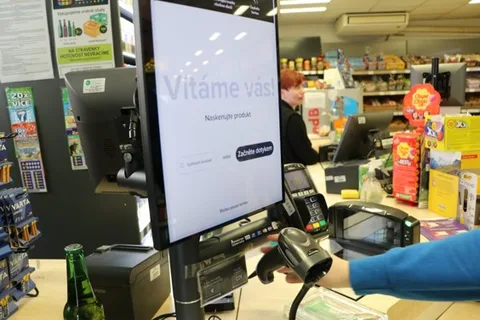 Detailní záběr na ruku zákazníka skenujícího produkt ručním skenerem u obrazovky samoobslužné pokladny.